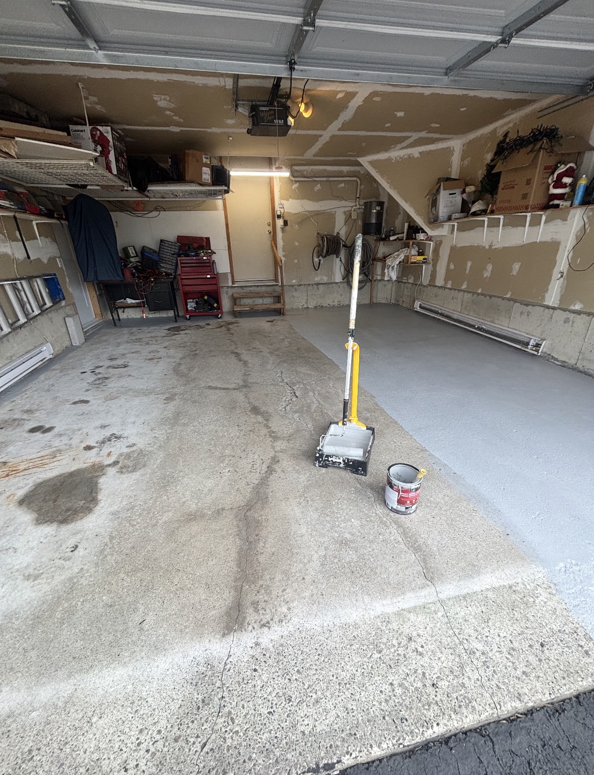 Garage floor being coated in light gray floor paint, with the untreated concrete still visible on the other half and a roller and open paint can in the foreground