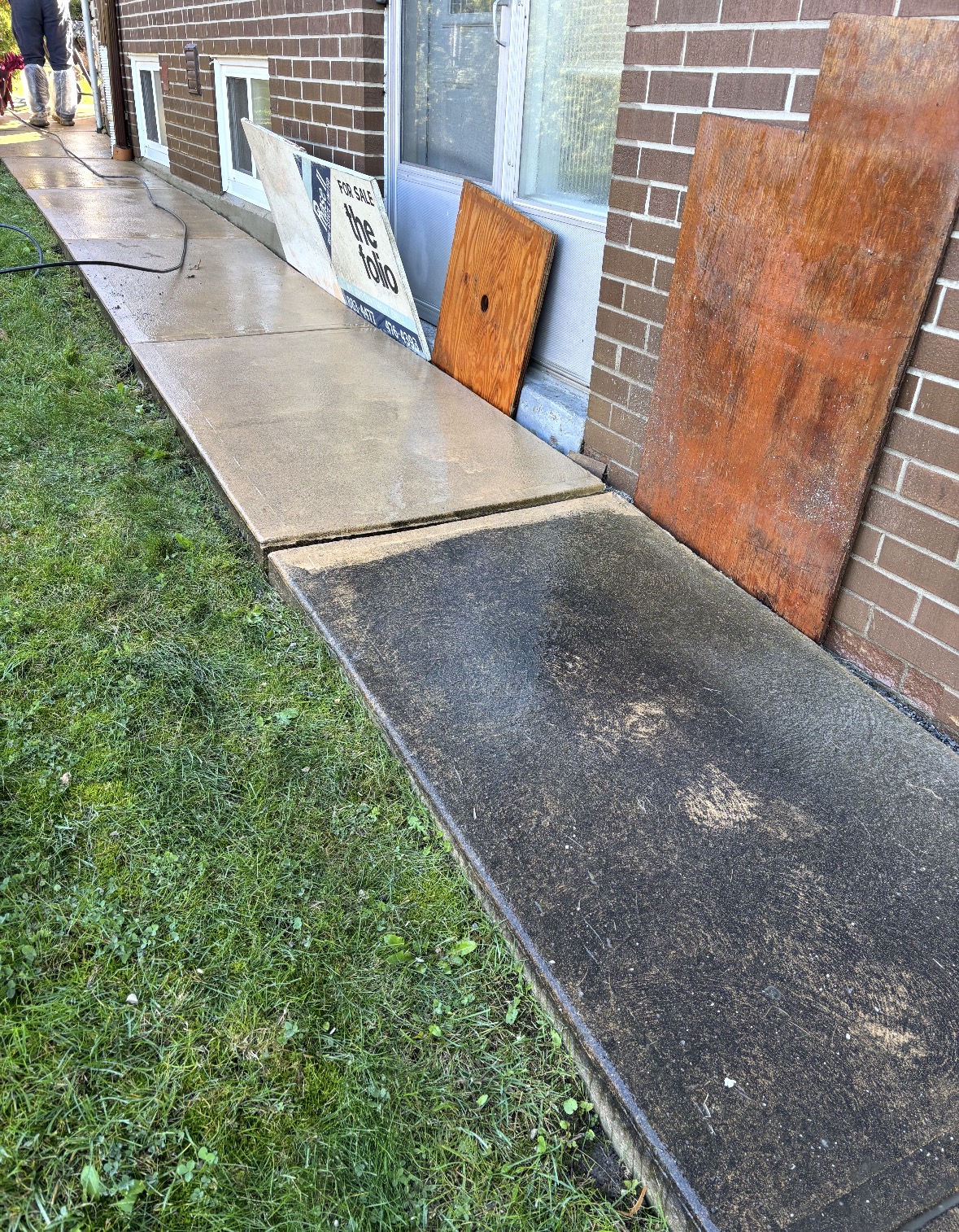 Concrete walkway beside a brick home, the nearest slab still dark with grime while the slabs beyond it are washed clean and wet, a lawn running along the left