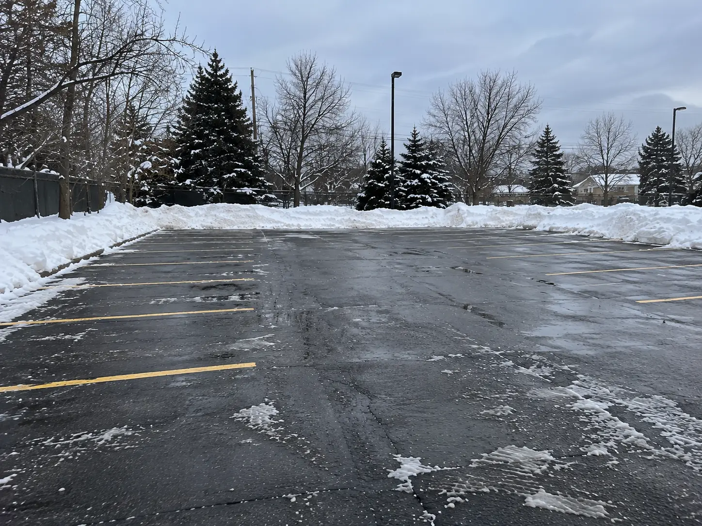 Cleared asphalt parking lot with painted yellow stall lines, thin slush and tire tracks on the pavement, and plowed snowbanks along the edges