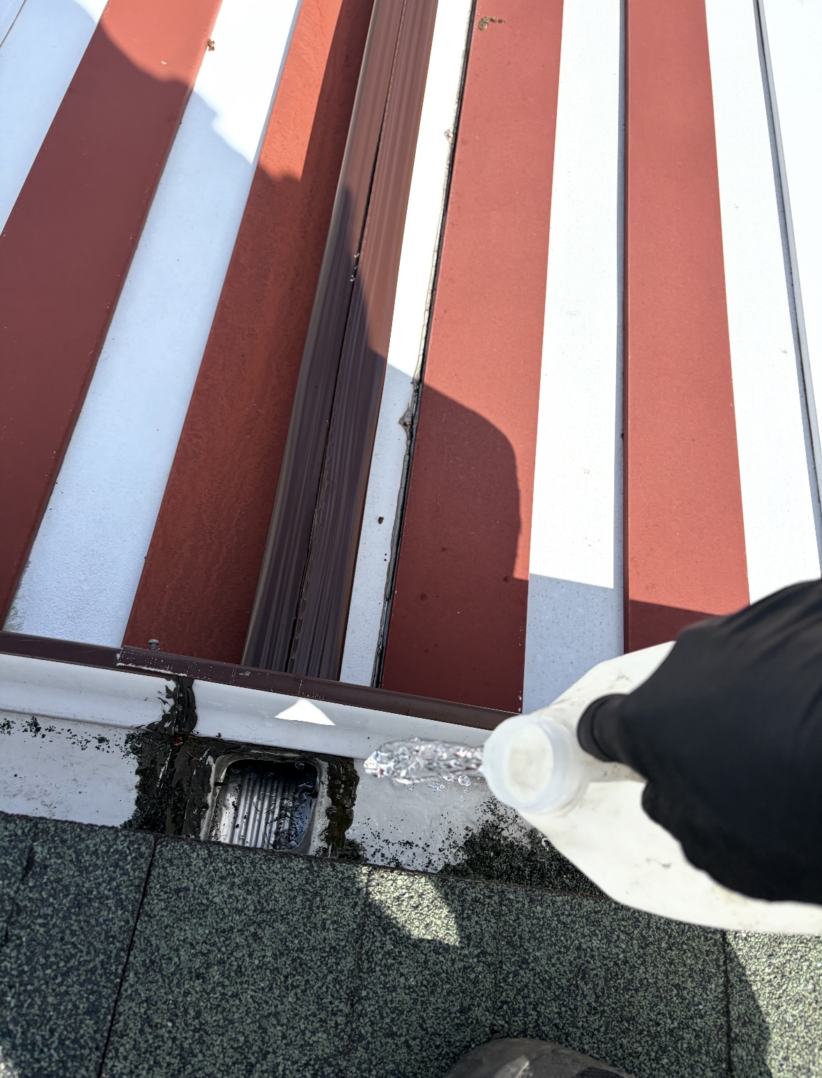 Gloved hand pouring water from a white jug into a gutter downspout opening beneath a red-and-white striped metal roof