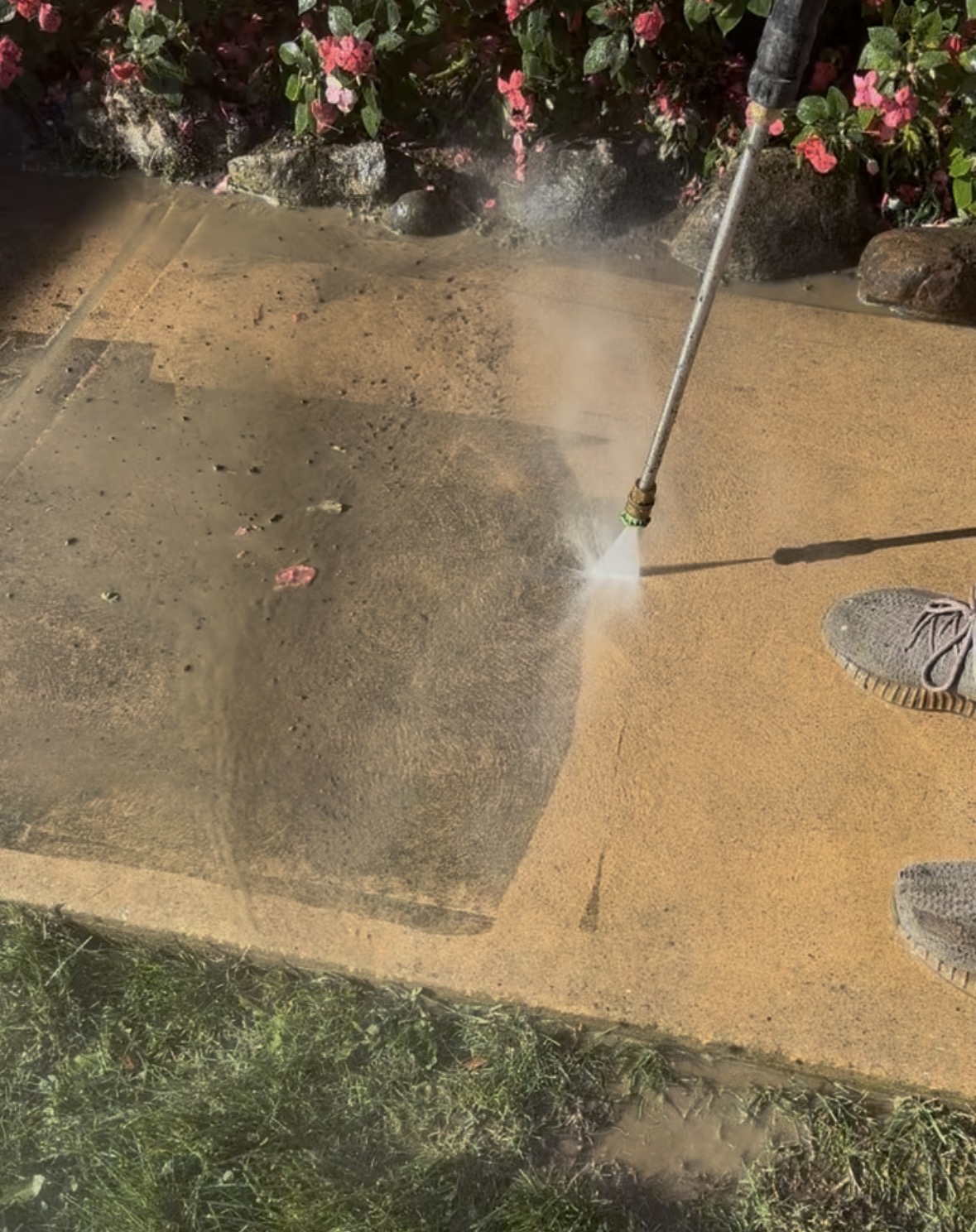 Pressure washer wand spraying a concrete walkway beside a flower bed, the cleaned right side a bright sandy tan against the dark, grimy left side, with green grass along the bottom