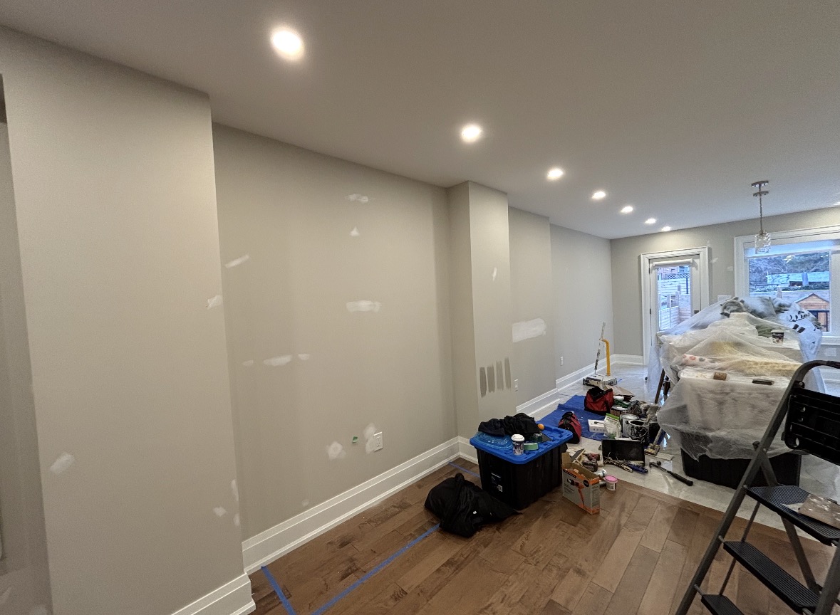 Open-concept living room prepped for painting, with spackled drywall, paint sample swatches on a wall column, furniture wrapped in plastic and a stepladder on the hardwood floor