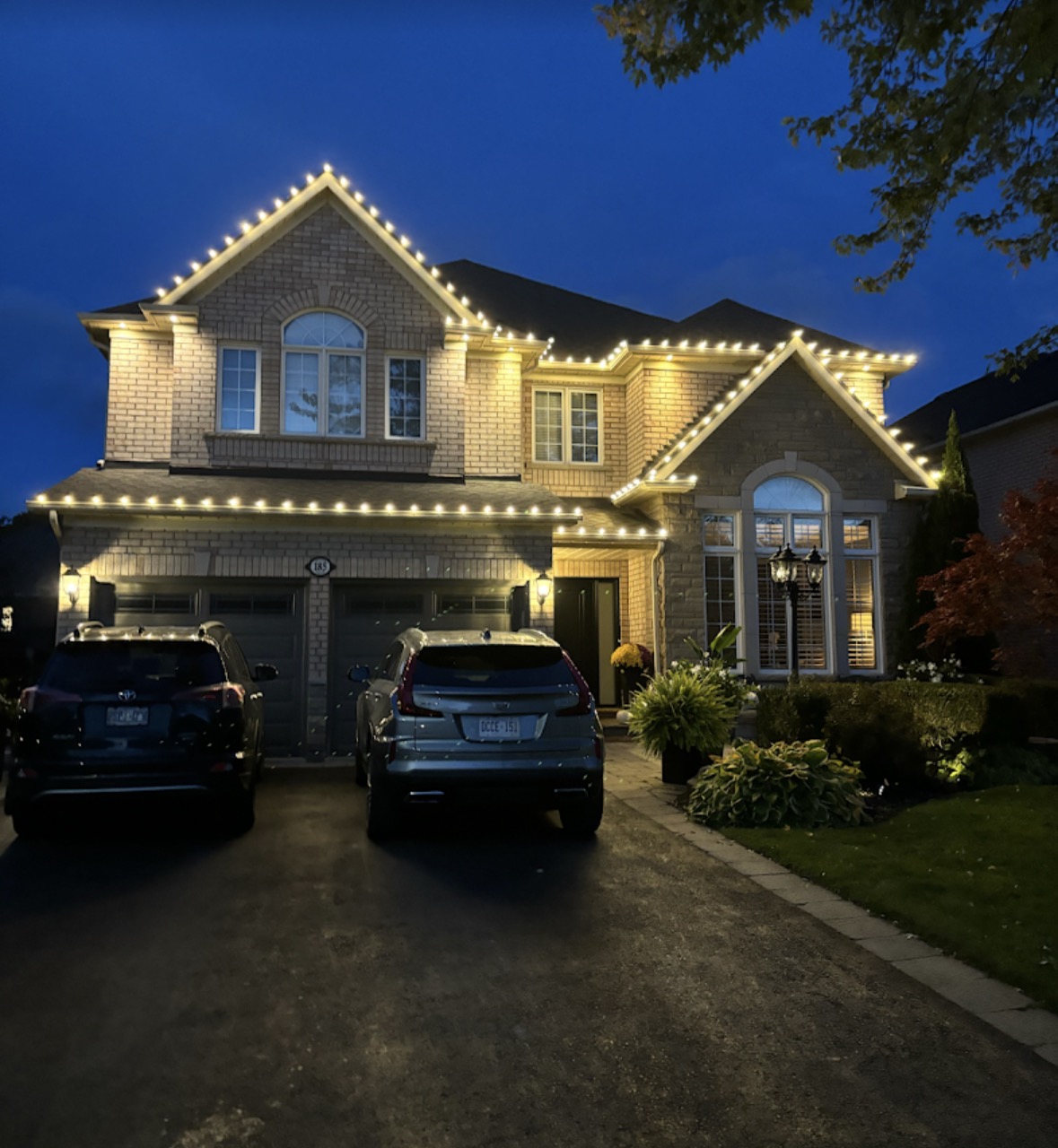 Two-storey brick home at dusk with warm-white holiday lights tracing the roofline, gables, and garage, with two cars in the driveway