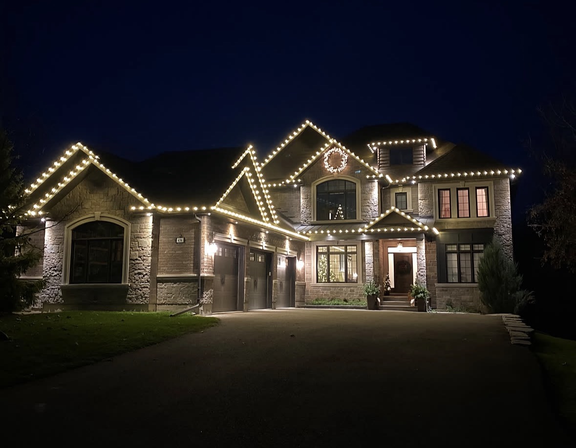 Stone home at night with warm-white lights outlining the roof gables and eaves, an illuminated wreath on the central gable, and a decorated Christmas tree visible through the front window
