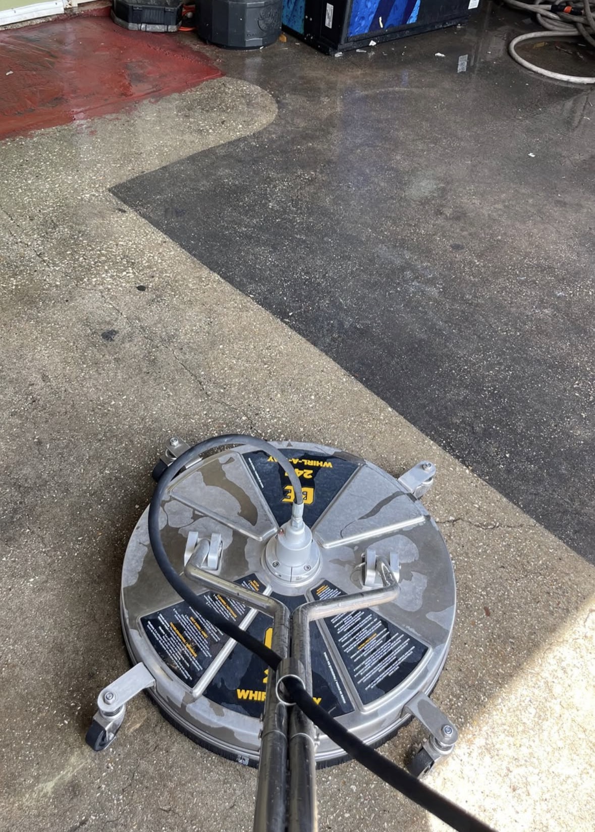 Round pressure washer surface cleaner resting on a concrete floor, the washed left side a light speckled tan against a sharp line of dark, uncleaned grime on the right