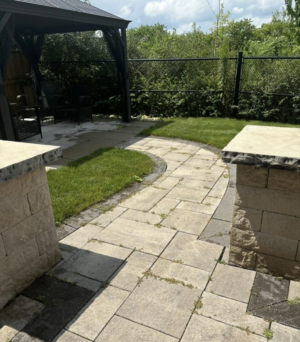 Weathered backyard paver walkway between two stone pillars, with weeds growing through the joints, before cleaning, sanding and sealing