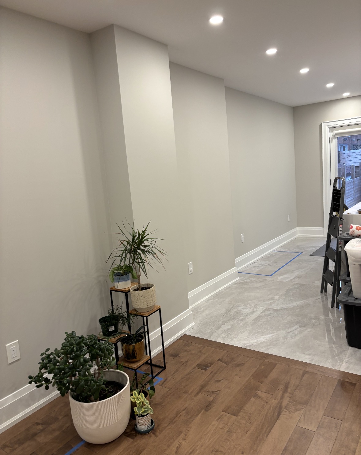 Prep work in a finished home interior, with blue layout tape marked on the wood and tile floors and a stepladder and bins staged near the walkout door