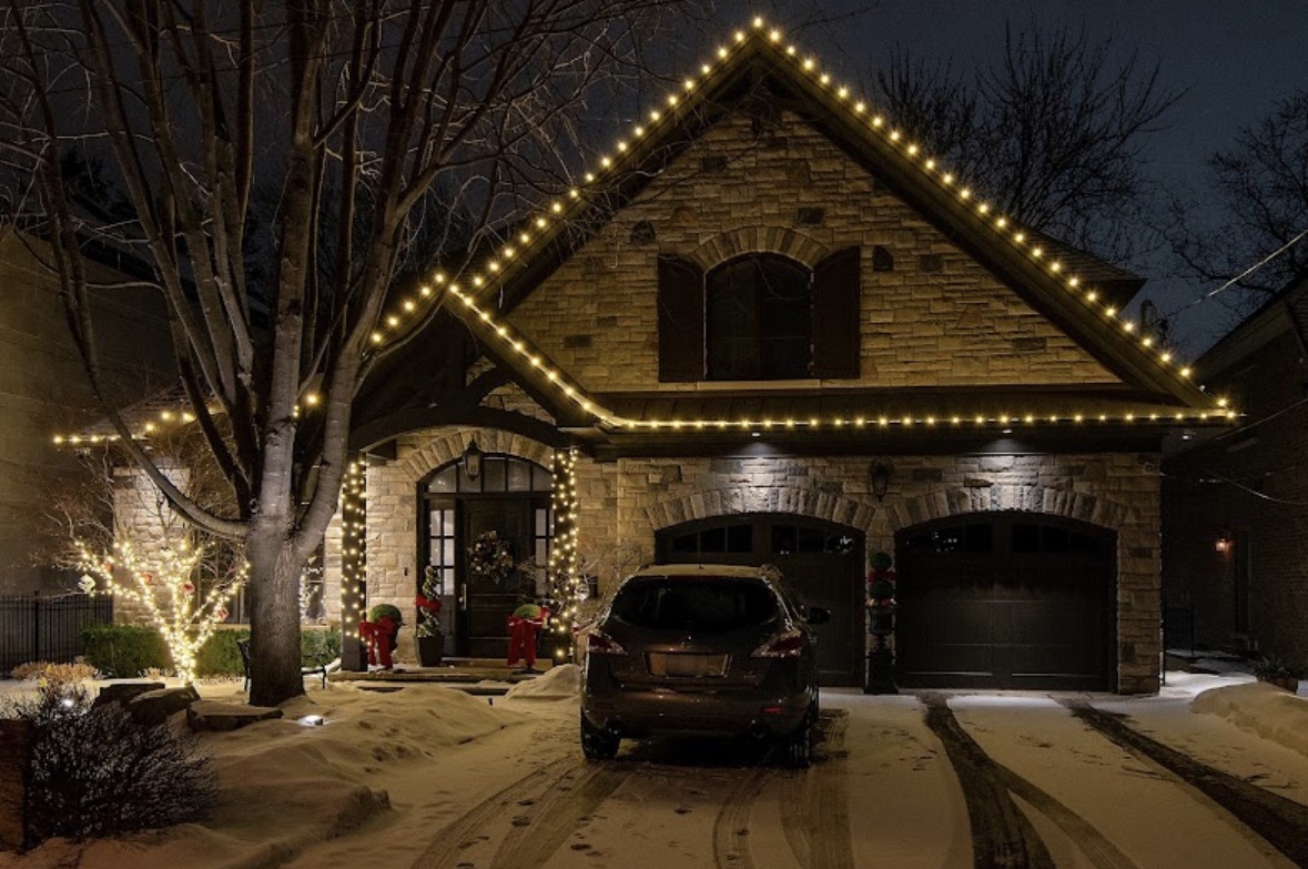 Two-storey home at night with warm-white lights along the roofline and gables, pillars wrapped at the front entrance, and a snow-covered driveway