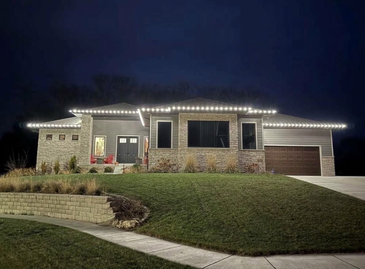 Single-storey home at night with warm-white lights along the rooflines above a stone-and-siding facade, a curved retaining wall and lawn in front