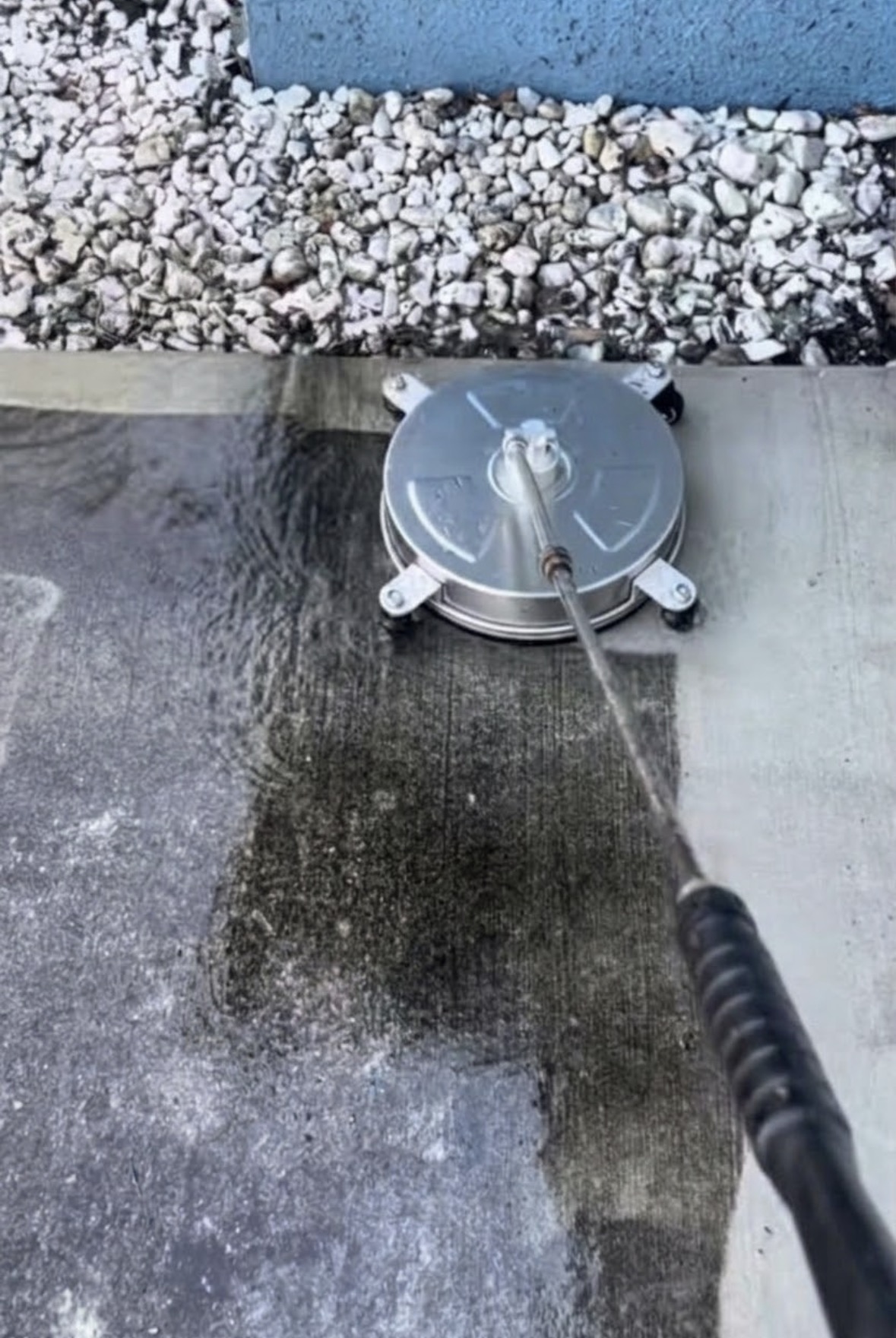 Concrete slab pressure washed from above, a round surface cleaner attachment resting on the wet pavement with the cleaned right side a bright light grey against the dark, grime-covered left